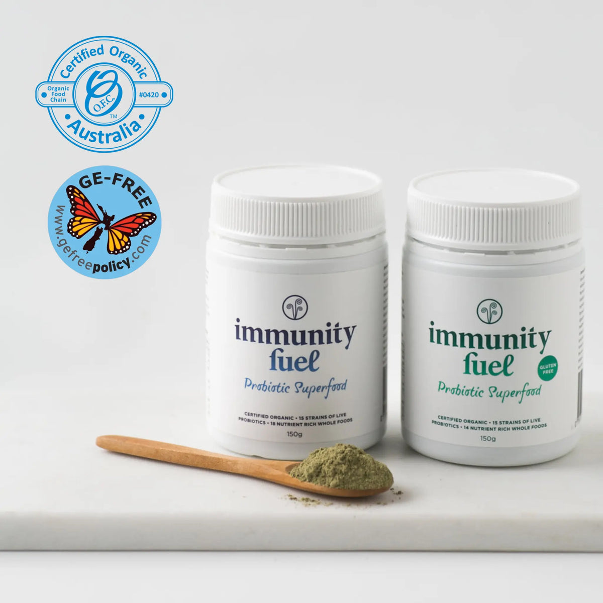 Two white plastic immunity fuel supplement jars on a white surface, with a small wooden spoon holding green powder in front. Two certification badges appear at the top left: Certified Organic Australia and GE-Free Policy.