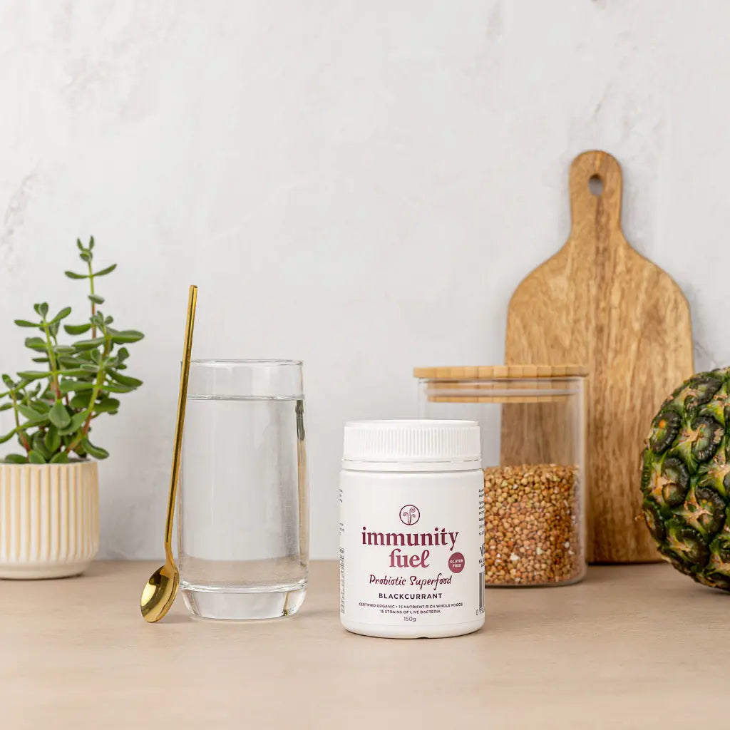 Jar of blackcurrant 'immunity fuel' probiotic prebiotic on a table with a glass of water, spoon, and pineapple.